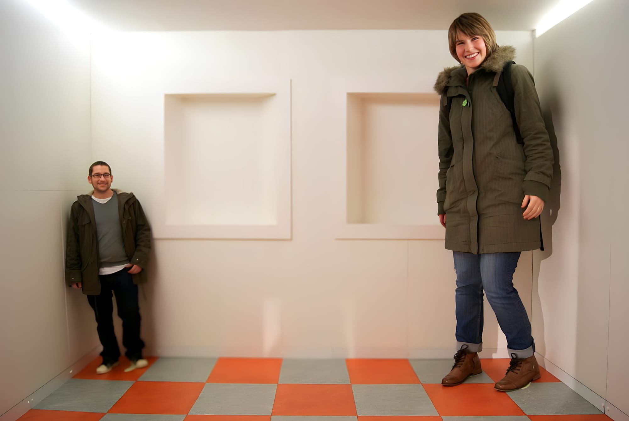 A photo of a man and a woman standing in the opposite sides of the room. The woman on the right looks much larger and taller than the man on the left, who looks smaller due to design of the Ames room. The Ames room is a specially designed room which demonstrates an optical illusion.