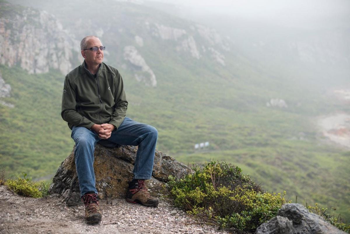 A picture of archaeologist Curtis Marean sitting on a rock, framed by scenic mountains in the background.