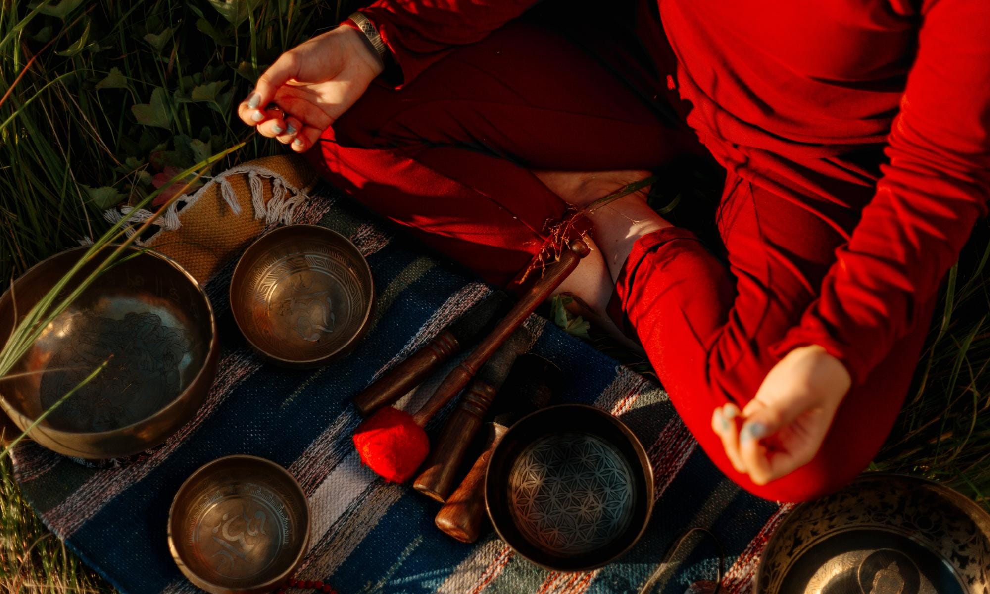  A photo of someone sat cross legged meditating outdoors, directed towards the ground where there are various bowls and tools. 