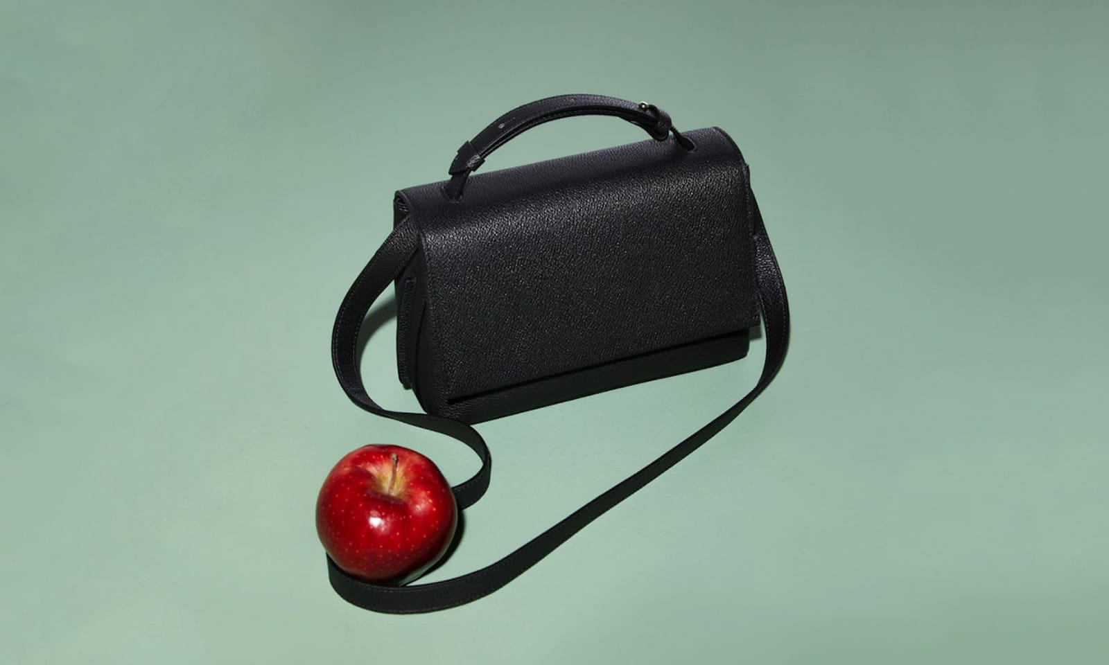 A photo of a black leather bag and a red apple against a light green background.